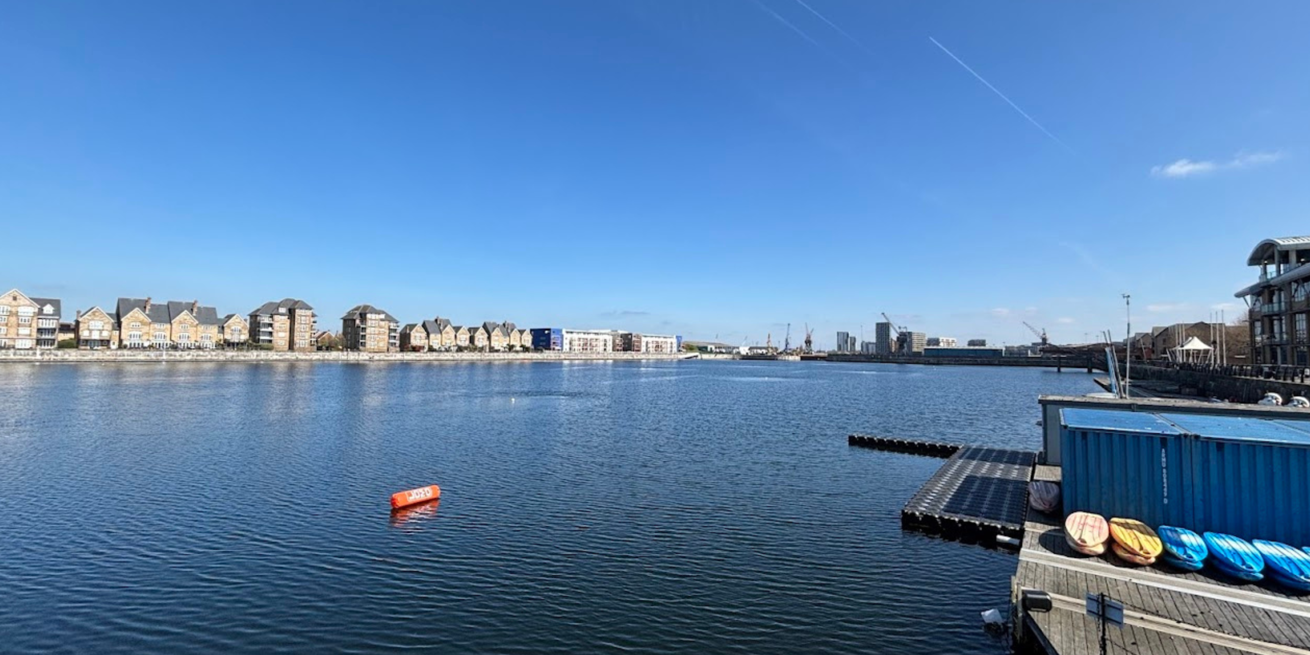 Waterfront with row of brick residential buildings along a calm harbor under a clear blue sky, with a dock and colorful kayaks on the right.