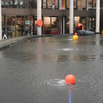 Urban canal street flooded; a small boat with people in high-visibility jackets afloat near storefronts, orange buoys drifting in the water.