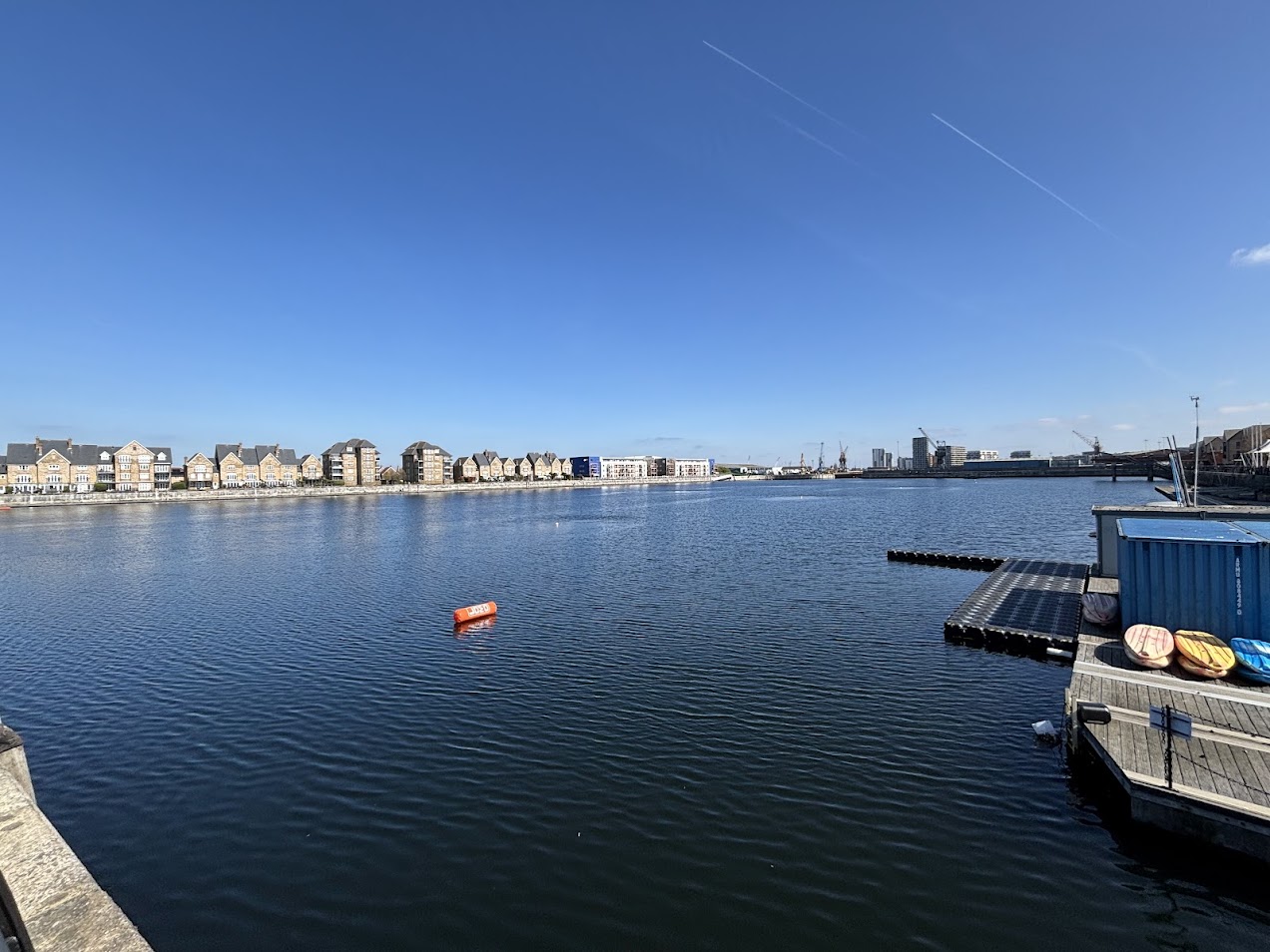 Calm harbor with row of townhouses along the water and a clear blue sky; an orange buoy floats near the center of the water.