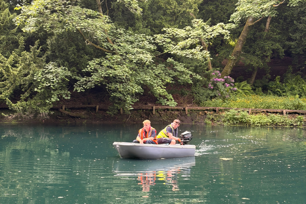 Two men in a small motorboat on a calm, greenish river, wearing orange and yellow life jackets beside a tree-lined bank. (informative)