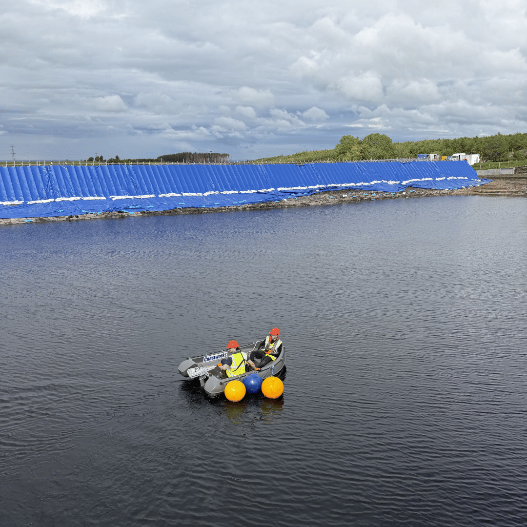 Two people in a small inflatable boat with orange helmets near a blue barrier along a water body.