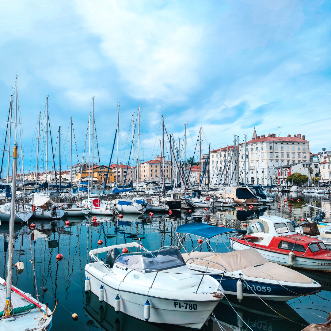 Harbor filled with moored sailboats and small boats, calm turquoise water reflecting masts, with pastel buildings along a sunny waterfront in the background.