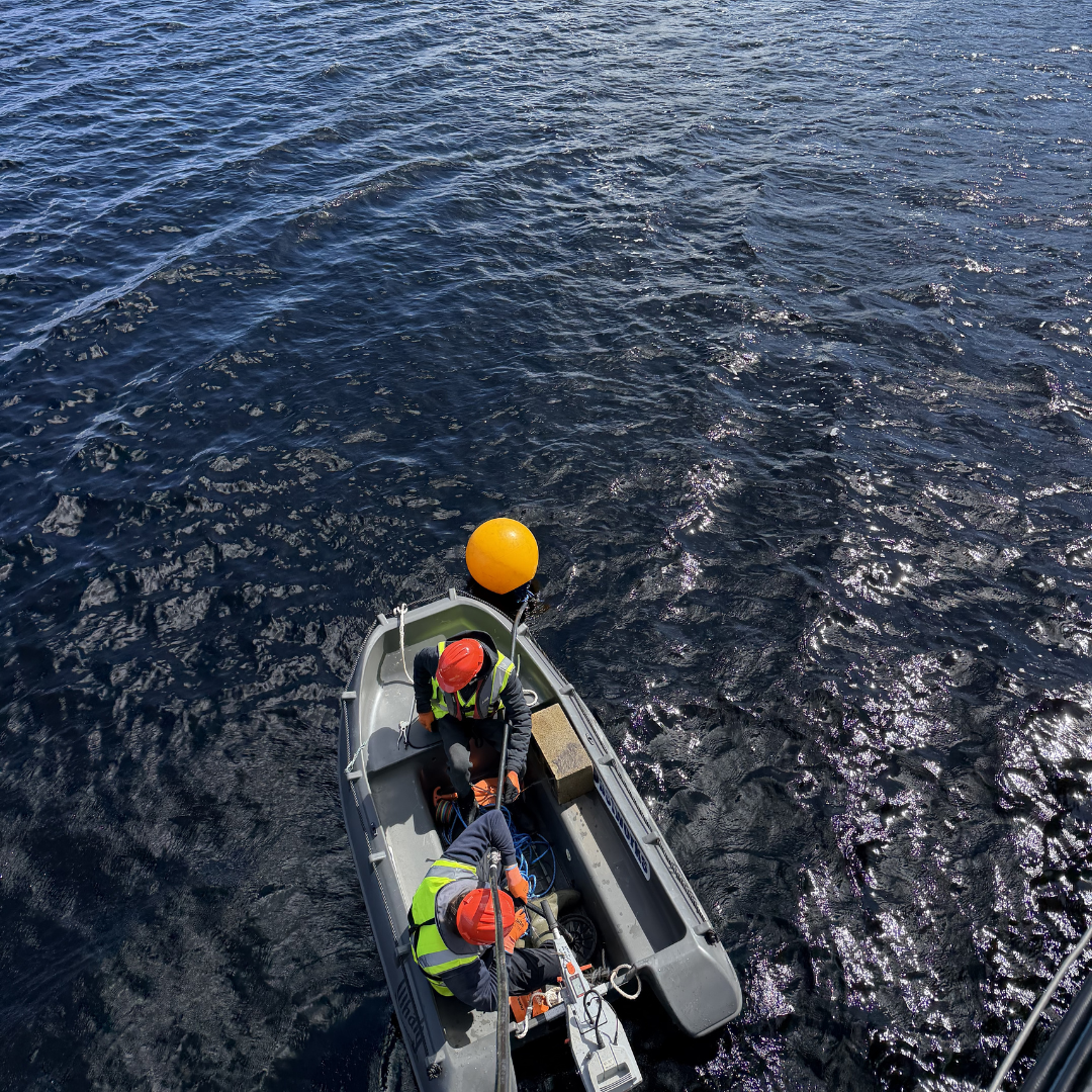 A small gray motorboat with two people in high-visibility vests and orange hard hats sits on dark blue water, tied to a bright orange buoy at the stern.